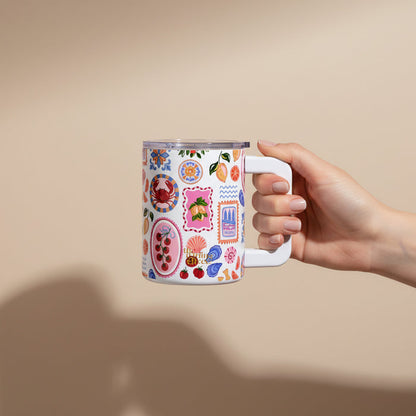 A hand holds The Darling Effect’s Ciao Bella 14 oz Insulated Mug, featuring colorful fruit and floral illustrations with decorative frames, set against a plain beige background.