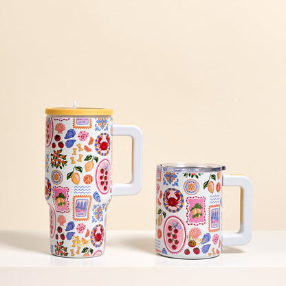 Two Ciao Bella 14 oz Insulated Mugs by The Darling Effect, each with a white handle, sit on a white surface. Their vibrant, playful designs include fruits, flowers, and geometric patterns in red, blue, pink, and yellow.