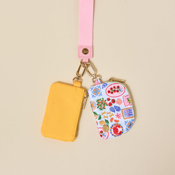 The Darling Effect’s Ciao Bella Dynamic Duo Pouch Wristlet features a pink lanyard with gold hardware and two pouches—one solid yellow, one white with fruit and floral patterns—for a vibrant, versatile set.