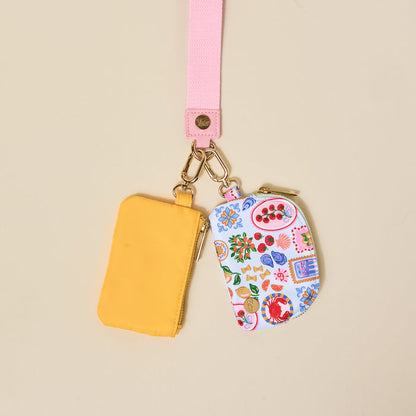 The Darling Effect’s Ciao Bella Dynamic Duo Pouch Wristlet features a pink lanyard with gold hardware and two pouches—one solid yellow, one white with fruit and floral patterns—for a vibrant, versatile set.
