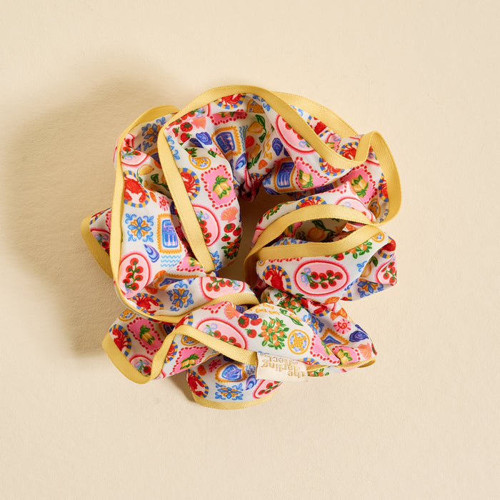 The Darling Effect's Ciao Bella Ruffle Up Scrunchie features a playful ruffled design with yellow trim and fruit illustrations, shown on a light beige background. Gentle on hair, it’s the perfect accessory to add fun to any hairstyle.