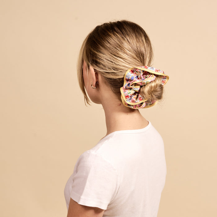 A woman with blonde hair in a low bun, secured with The Darling Effect's Ciao Bella Ruffle Up Scrunchie. She wears a white t-shirt and faces away from the camera against a plain beige background.