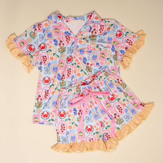 The Darling Effect’s Ciao Bella Slumber Party Pajama Shorts Set features a light blue, ultra-soft fabric with playful crabs, fruit, flowers, and postcards. This relaxed fit has a ruffled gold-trimmed button-up top and shorts with pink accents.