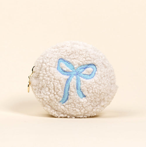 The Circle Bow Blue Teddy Pouch by the-darling-effect is a round, white sherpa pouch with a light blue embroidered bow on the front and a small gold zipper on the side, shown against a plain light background.