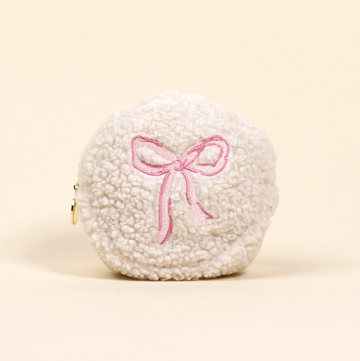 The Circle Bow Pink Teddy Pouch by the-darling-effect, made from soft white sherpa with a pink embroidered bow and gold zipper pull, is displayed on a plain beige background.