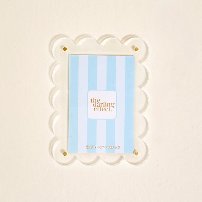 A clear scalloped acrylic photo frame magnet by The Darling Effect features gold corner screws and holds a blue and white striped card. The Clear Mini Acrylic Picture Frame Magnet is shown against a plain off-white background.