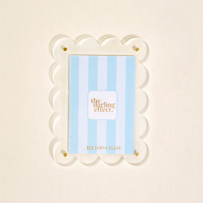A clear scalloped acrylic photo frame magnet by The Darling Effect features gold corner screws and holds a blue and white striped card. The Clear Mini Acrylic Picture Frame Magnet is shown against a plain off-white background.