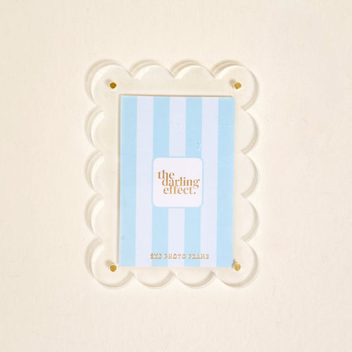 A clear scalloped acrylic photo frame magnet by The Darling Effect features gold corner screws and holds a blue and white striped card. The Clear Mini Acrylic Picture Frame Magnet is shown against a plain off-white background.