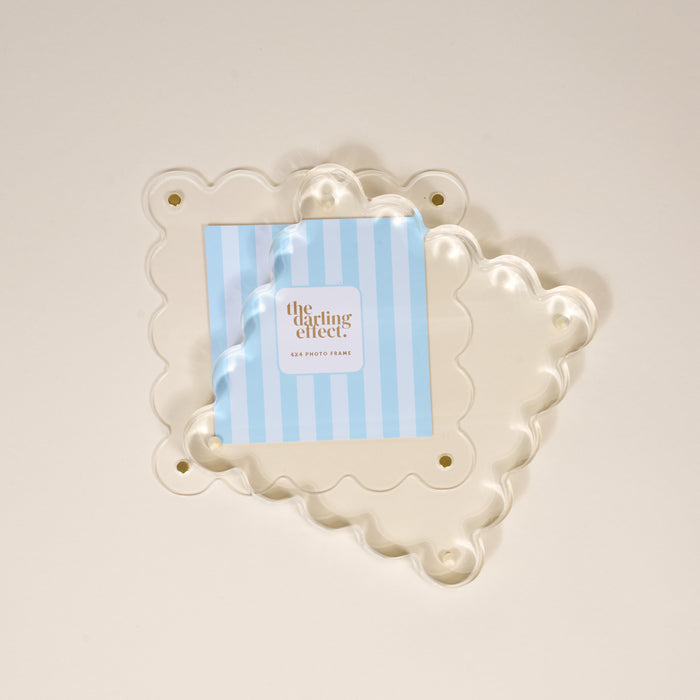 A Clear Square Scalloped Acrylic Frame by the-darling-effect is displayed over a cream background, holding a blue and white striped card with the brand name. This modern 6x6 photo frame features scalloped edges and four corner holes.