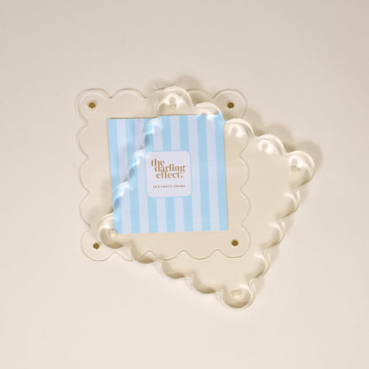A Clear Square Scalloped Acrylic Frame by the-darling-effect is displayed over a cream background, holding a blue and white striped card with the brand name. This modern 6x6 photo frame features scalloped edges and four corner holes.
