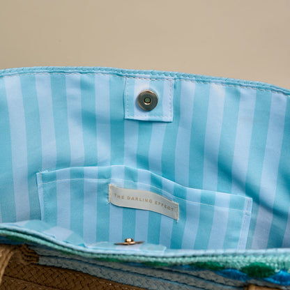 The Darling Effect's Coastal Breeze Straw & Sun Tote features an open design, light blue and white striped lining, an interior pocket, brand label inside, and a magnetic snap closure for easy access to your essentials.