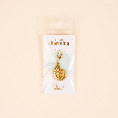 The Darling Effect's Conch Shell Jewelry Charm Gold features a clasp and comes on a white card reading "OH SO charming" and The Darling Effect, packaged in a clear bag with a light beige background—ideal for your charm bar or unique jewelry.
