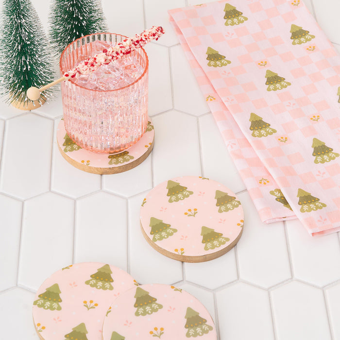 A pink glass with a festive stir stick sits on a round coaster with green Christmas trees. Two more coasters and The Darling Effect’s Countryside Christmas Tea Towel enhance the holiday décor on the white hexagon tile surface.