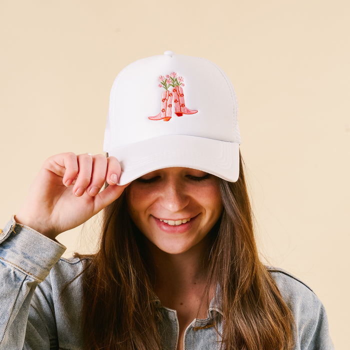 A smiling person touches the brim of the-darling-effect's Cowgirl Boots Trucker Hat, a trendy white cap featuring pink boots with flowers, against a plain beige background.