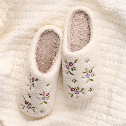 The Darling Effect's Dainty Floral Purple Fuzzy Slippers rest on a textured cream-colored blanket, showcasing their white plush design and purple and green floral accents.
