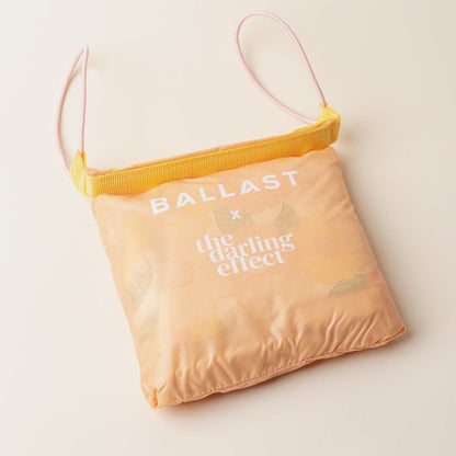 The Daisy Craze Peach Ballast Beach Pillow by The Darling Effect, featuring a peach floral pattern, yellow zipper, and pink strap, sits on a neutral surface—highlighting this limited-edition collaborative design.