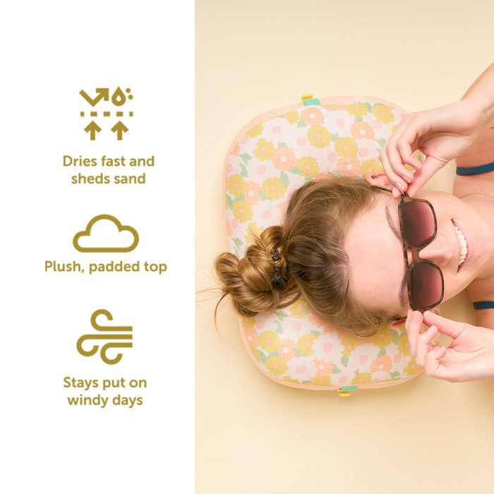 A woman relaxes on The Darling Effect’s Daisy Craze Peach Ballast Beach Pillow, smiling as icons highlight: “Dries fast and sheds sand,” “Plush, padded top,” and “Stays put on windy days.”.