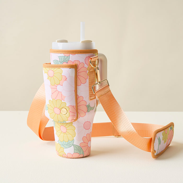 The Darling Effect’s Daisy Craze Peach Tumbler Sling, a floral-patterned tumbler with straw, sits in its matching neoprene carrier featuring an adjustable strap and side pocket, displayed on a light surface against a neutral background.