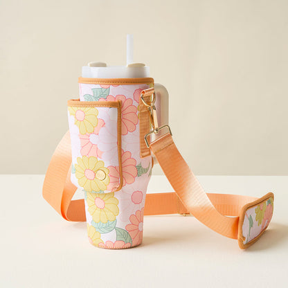The Darling Effect’s Daisy Craze Peach Tumbler Sling, a floral-patterned tumbler with straw, sits in its matching neoprene carrier featuring an adjustable strap and side pocket, displayed on a light surface against a neutral background.