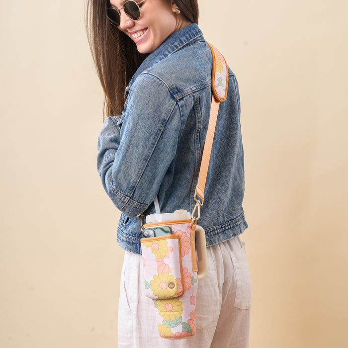 A woman in sunglasses, a denim jacket, and white pants smiles while carrying The Darling Effect’s Daisy Craze Peach Tumbler Sling, an adjustable floral-patterned carrier that holds her water bottle and phone.
