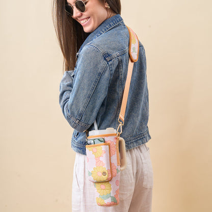 A woman in sunglasses, a denim jacket, and white pants smiles while carrying The Darling Effect’s Daisy Craze Peach Tumbler Sling, an adjustable floral-patterned carrier that holds her water bottle and phone.