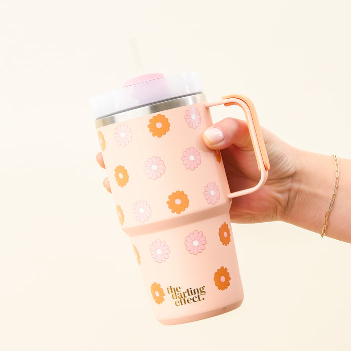 A hand holds The Darling Effect's Darling Daisy Peach 20 oz Mini On-The-Go tumbler with a handle, straw, and orange-pink flower patterns. "the darling effect" is printed on this insulated tumbler against a light beige background.