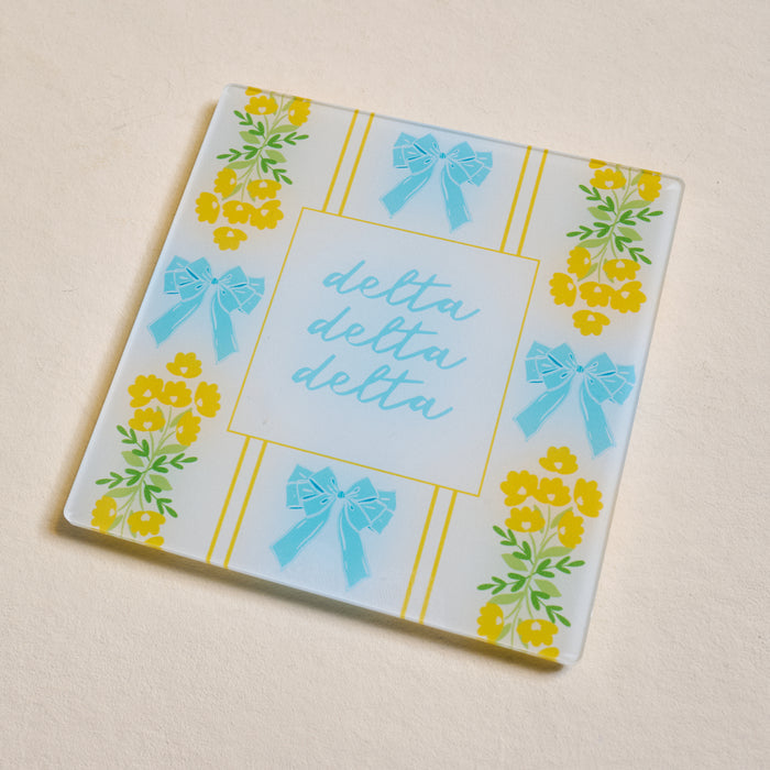 The Darling Effect Delta Delta Delta Acrylic Coaster features blue bows and yellow flowers bordering "delta delta delta" in blue cursive—perfect for Big/Little gifts or to display your sorority pride.