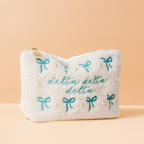 The Darling Effect’s Delta Delta Delta Bows & Pearls Teddy Pouch is a perfect sorority gift, featuring blue embroidered bows, faux pearls, teal script, and a peach-accented white sherpa design.