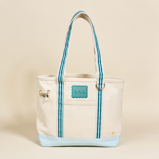 The Delta Delta Delta Canvas Tote Bag by The Darling Effect has cream canvas, blue/white striped handles, a blue wave patch, a gold pin with small charms on the left, and a light blue bottom—perfect for your sorority style.