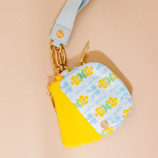 The Darling Effect's Delta Delta Delta Dynamic Duo Pouch Wristlet features a blue wrist strap, gold hardware, a bright yellow pocket, and a floral blue pocket with “Delta Delta” text and yellow flowers on a beige background.