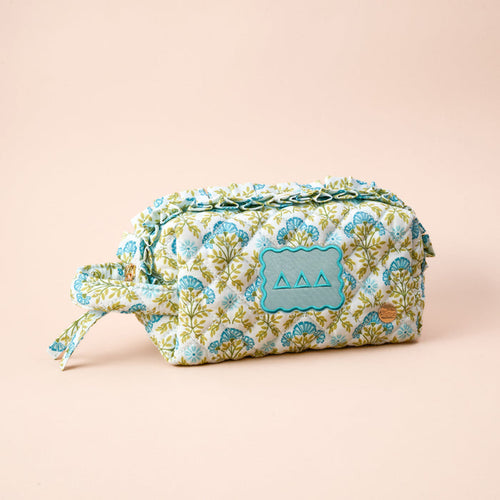 The Darling Effect's Delta Delta Delta Essentials Quilted Cosmetic Pouch features a blue and green floral pattern, a front blue patch with three triangles, and a zipper, shown resting on a light beige background.