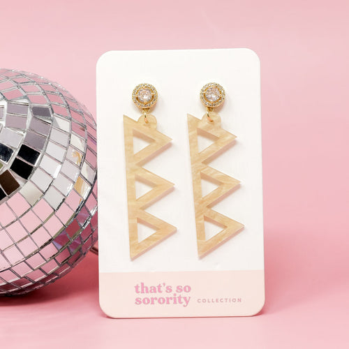 The Darling Effect's Delta Delta Delta Letter Gold Acrylic Earrings, displayed on a white card next to a disco ball against a pink backdrop—perfect for adding sparkle to any bid-day basket.