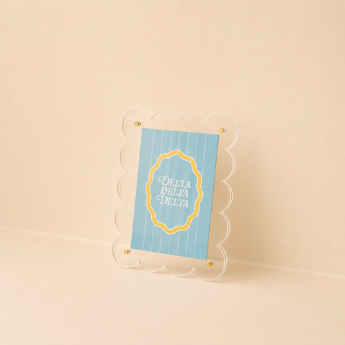 A blue and yellow “Delta Delta Delta” card is shown in The Darling Effect’s Mini Acrylic Picture Frame Magnet, featuring a scalloped clear acrylic design, set on a beige surface with a matching beige background.