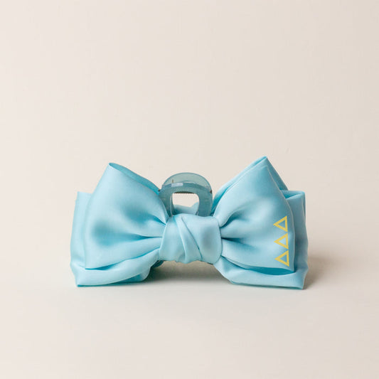 The Darling Effect's Delta Delta Delta Satin Bow Claw Clip features a light blue satin bow on a translucent blue clip with three small yellow triangle symbols, adding elegant charm to updos against a plain light background.