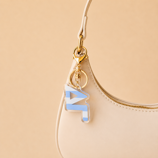 A cream-colored handbag with a gold zipper, featuring The Darling Effect’s Delta Gamma Stripes Acrylic Keychain in blue and white—a perfect sorority gift for bid day—set against a beige background.