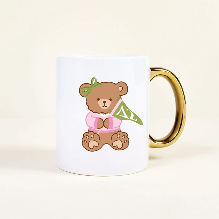 The Darling Effect’s Delta Zeta 11 oz Cozy Cute Ceramic Mug features a brown bear in a pink shirt with a green bow, holding a green pennant with white ΔΖ letters—perfect for sorority sisters or as a charming gift.