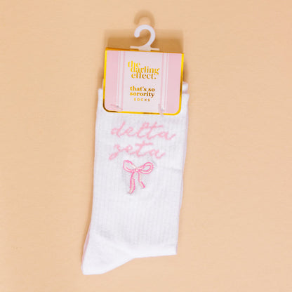 Delta Zeta Embroidered Crew Socks by The Darling Effect feature white ribbed fabric, pink "delta zeta" embroidery, and a bow—packaged for gifting and perfect as cute sorority gifts.
