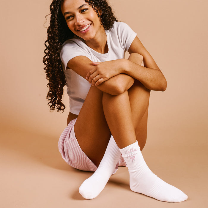 A young woman with curly hair smiles, sitting cross-legged and wearing The Darling Effect's Delta Zeta Embroidered Crew Socks—an ideal sorority gift.