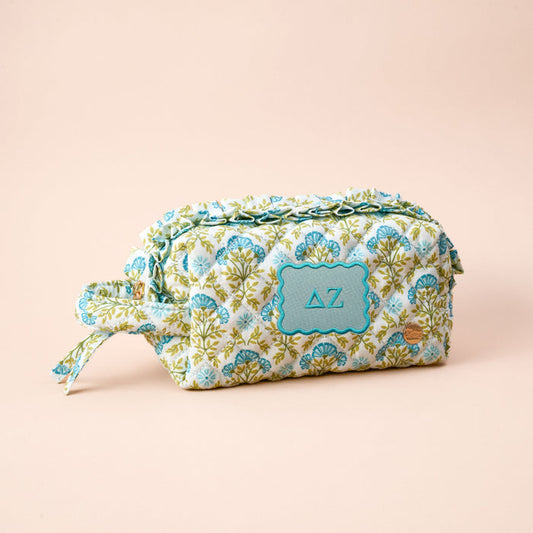 The Darling Effect’s Delta Zeta Essentials Quilted Cosmetic Pouch features a floral design, zipper closure, and teal label with Greek letters—an essential for any sorority collection. Shown on a plain beige background.