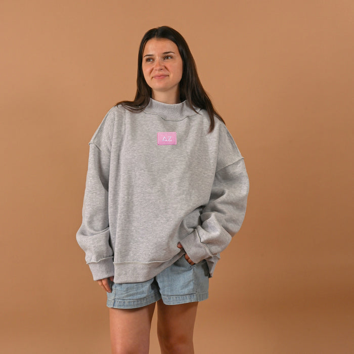 A woman smiles softly with one hand in her pocket, wearing The Darling Effect’s Delta Zeta Letter Patch Mockneck Sweatshirt (S/M) and light denim shorts, posed against a plain brown background.