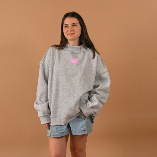 A woman smiles softly with one hand in her pocket, wearing The Darling Effect’s Delta Zeta Letter Patch Mockneck Sweatshirt (S/M) and light denim shorts, posed against a plain brown background.