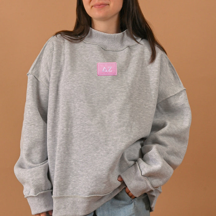 A person wearing an oversized gray Delta Zeta Letter Patch Mockneck Sweatshirt by The Darling Effect, featuring a pink patch with white Greek letters on the front, stands against a plain tan background—perfect for showing off your sisterhood style.