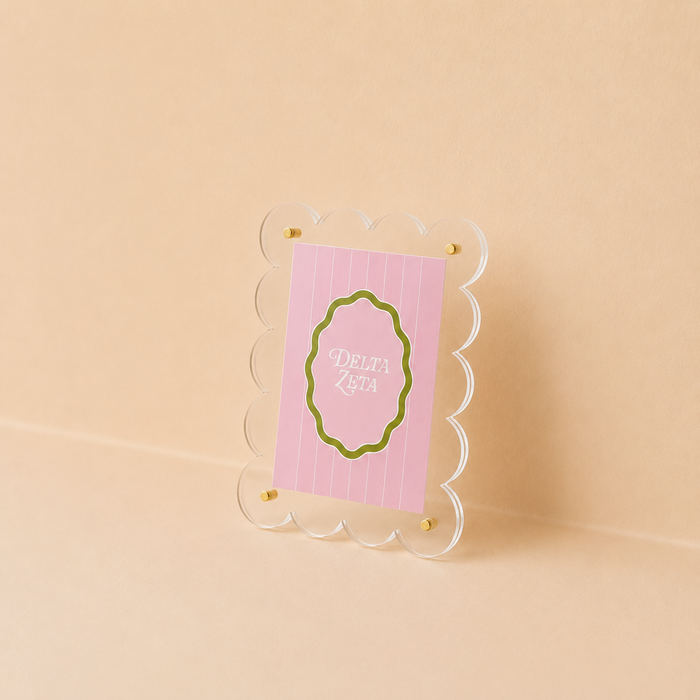 The Darling Effect Delta Zeta Mini Acrylic Picture Frame Magnet features a pink card with "Delta Zeta" in white text inside a green oval, accented by gold fasteners and displayed in a clear scalloped acrylic frame against a light beige background.