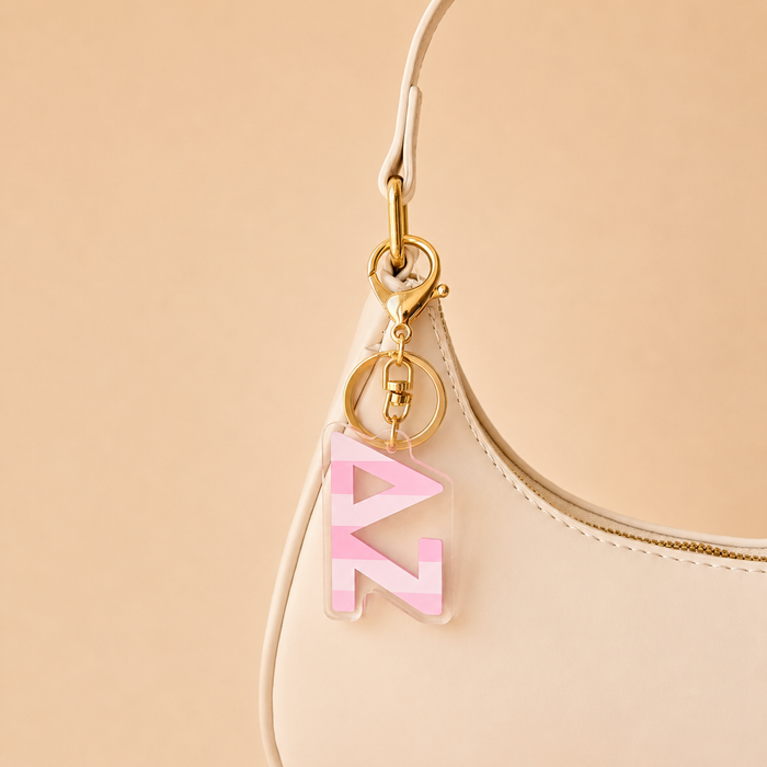 A cream-colored handbag with a gold keychain on the strap, featuring The Darling Effect’s Delta Zeta Stripes Acrylic Keychain—perfect as a chic sorority gift or bid day accessory against a beige background.