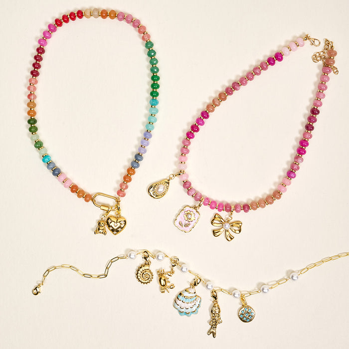 Three colorful beaded necklaces from The Darling Effect feature gold jewelry charms, including the Diamond Heart Jewelry Charm Gold, a bow, cupcake, fish, sun, and floral motifs. One necklace also has gold links and pearls on a light background.