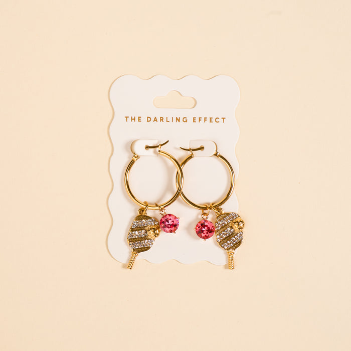The Darling Effect’s Dinking Around Signature Hoop Earrings feature gold hoops with pickleball paddle and pink gemstone charms, adding playful sparkle to your everyday style.