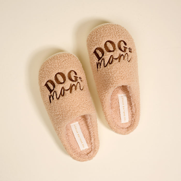 The Darling Effect's Dog Mom Fuzzy Slippers are plush beige slippers featuring "DOG mom" and a paw print embroidery, shown resting on a light-colored surface.