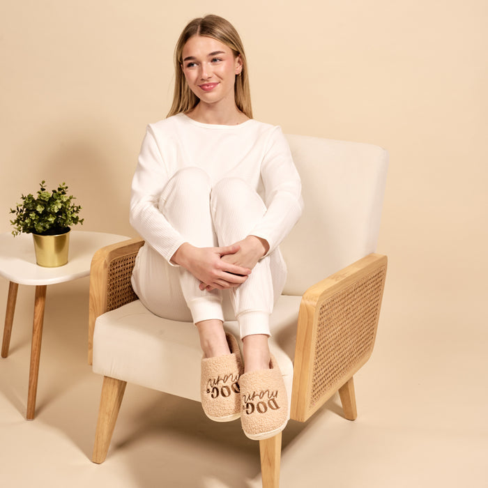 A young woman in white loungewear smiles as she sits on a beige armchair, wearing The Darling Effect’s Dog Mom Fuzzy Slippers. A small potted plant rests on a table nearby, with a light beige background completing the cozy scene.