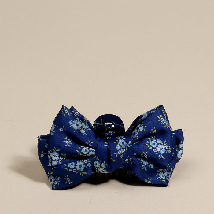 A blue bow tie with small light blue flowers and green leaves sits on a beige background—beautifully coordinated with The Darling Effect’s Endless Daydream Navy Mini Satin Bow Claw Clip.