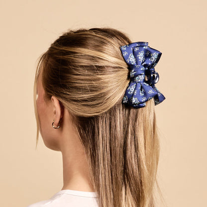 A woman with straight blonde hair styled half-up, half-down secures it with The Darling Effect’s Endless Daydream Navy Mini Satin Bow Claw Clip, which features a blue and white floral pattern. She is seen from behind against a beige background.
